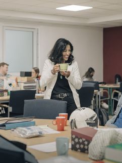 Atika Müller-Eroğul im Lehrerzimmer mit einer Tasse in der Hand und im Gespräch mit einem Kollegen, der eine Mund-Nasen-Bedeckung trägt und auf einem Stuhl sitzt.