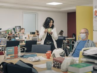 Atika Müller-Eroğul im Lehrerzimmer mit einer Tasse in der Hand und im Gespräch mit einem Kollegen, der eine Mund-Nasen-Bedeckung trägt und auf einem Stuhl sitzt.