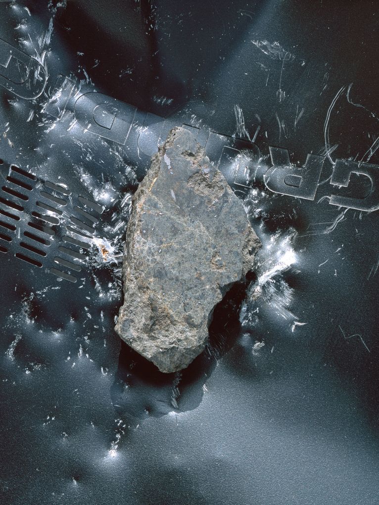 Grauer Meteorit, der auf das Metallgehäuse ein Geräts der Marke Grundig aufgetroffen ist und deutliche Spuren hinterlassen hat.