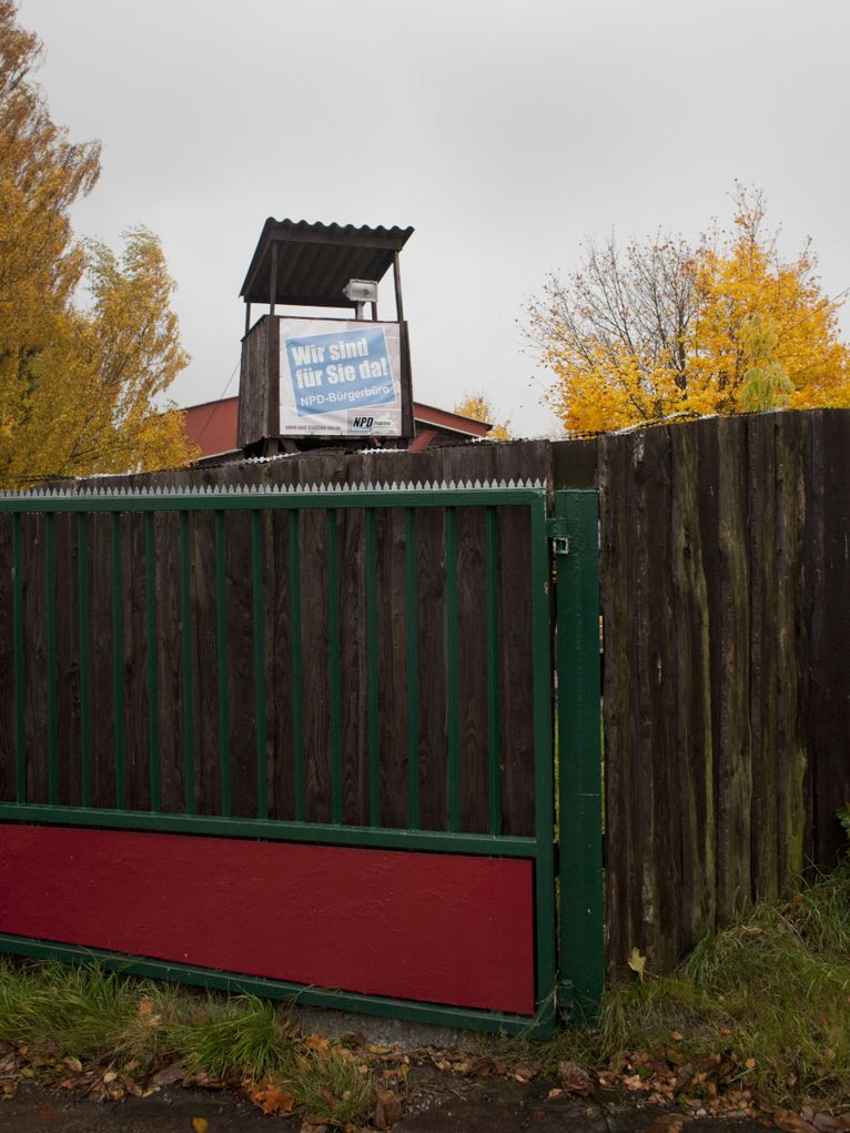 Wachturm aus Holz, auf dem ein NPD-Plakat klebt, hinter einem hohen Gatter.