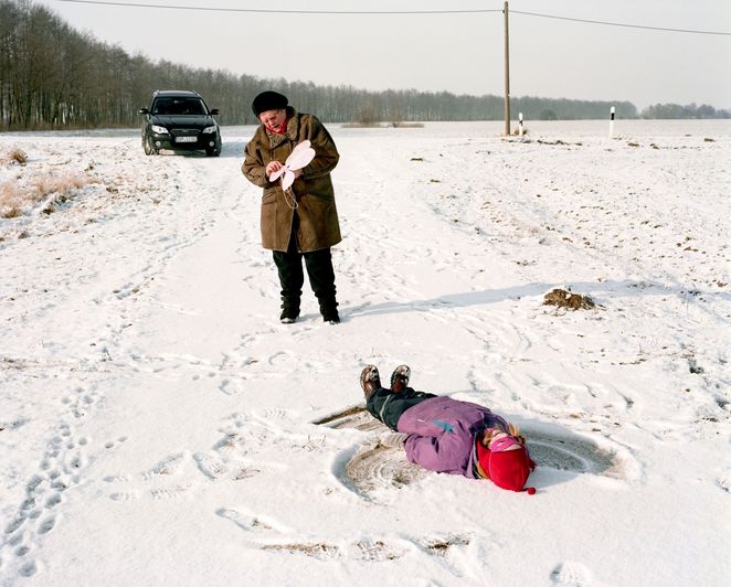 Eine ältere Frau und ein Kind im Schnee, das einen Schneeengel macht. Im Hintergrund ein schwarzes Auto vor einem Waldrand.