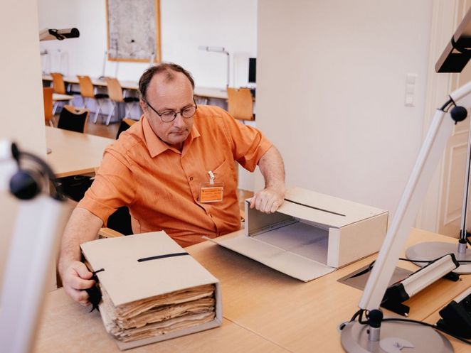 Kreuter arbeitet an einem Tisch in einem Archiv- oder Lesesaal. Vor ihm liegt eine geöffnete Archivbox mit historischen Dokumenten, daneben ein weiterer Archivkarton.