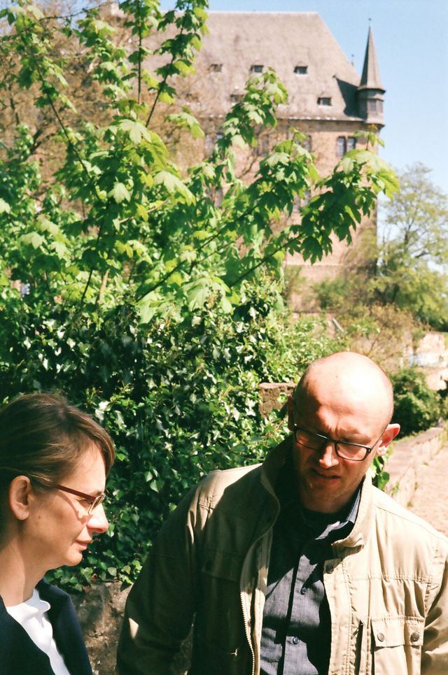 Ksenia Stanicka-Brzezicki und SŁAWEK Brzezicki vor einem grünen Busch, im Hintergrund ein großes Gebäude.