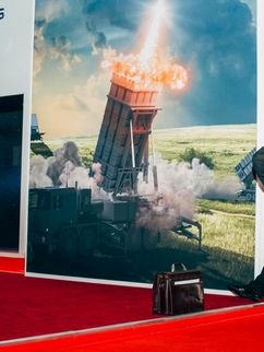 Zwei Herren in Anzügen, einer in Uniform for einem Plakat einer Raketenabwehr-Maschine.zu sehen und einer in Uniform, der ebenfalls nur halb zu sehen ist, von
