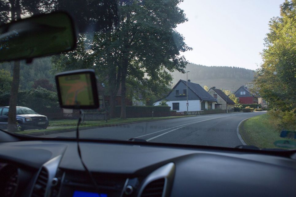 Blick aus der Frontscheibe eines Autos auf eine Straße, Bäume und einige Häuser.