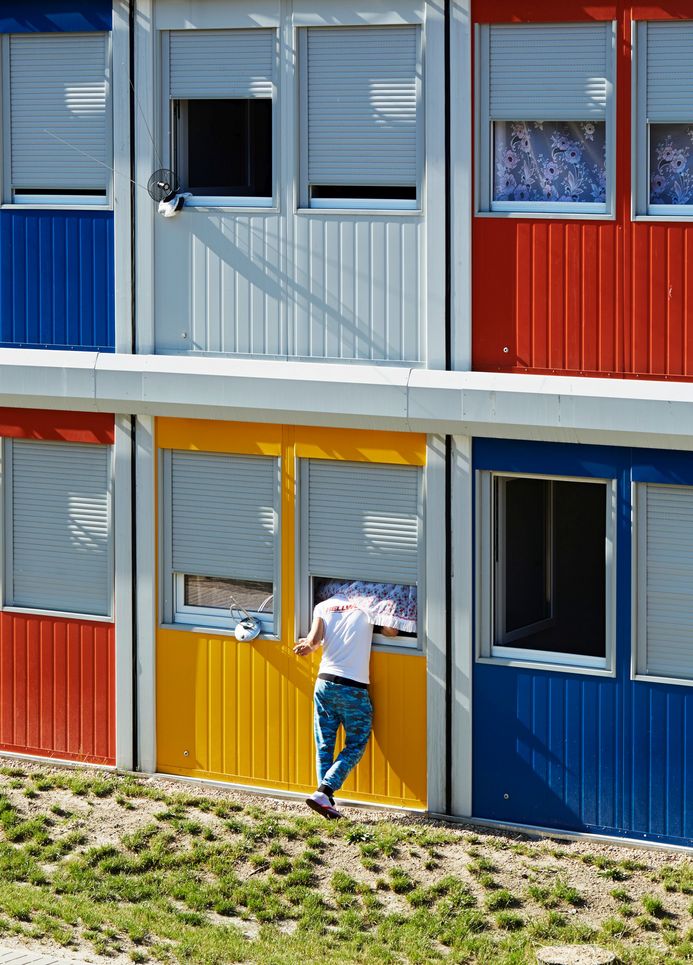 Bunte, übereinander gestapelte Wohncontainer. In ein Fenster steckt ein Mann seinen Kopf hinein, er wird von einem Vorhang verdeckt.