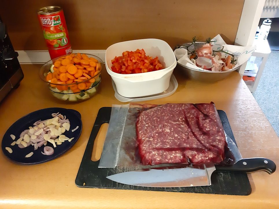 Fleisch auf einem Brett, ein großes Messer daneben. Geschnittenes Gemüse in Schüsseln.