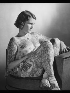Die am ganzen Körper tätowierte Lady Betty Broadbent sitzt mit überschlagenen Beinen auf einem Sessel.