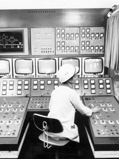 Kraftwerk Vockerode In einem Kontrollraum sitzt eine Person vor einem großen Bedienpult mit zahlreichen Schaltern, Anzeigen und Bildschirmen. Die Schwarzweißaufnahme zeigt einen technischen Schaltraum in einem Industriebetrieb.