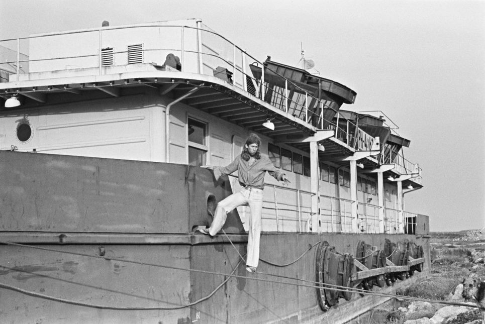 Schwarzweißaufnahme eines Schiffes mitten in der Wiese, an dem sich ein Mann mit langen Haaren seitlich festhält, der auf einem Seil steht.