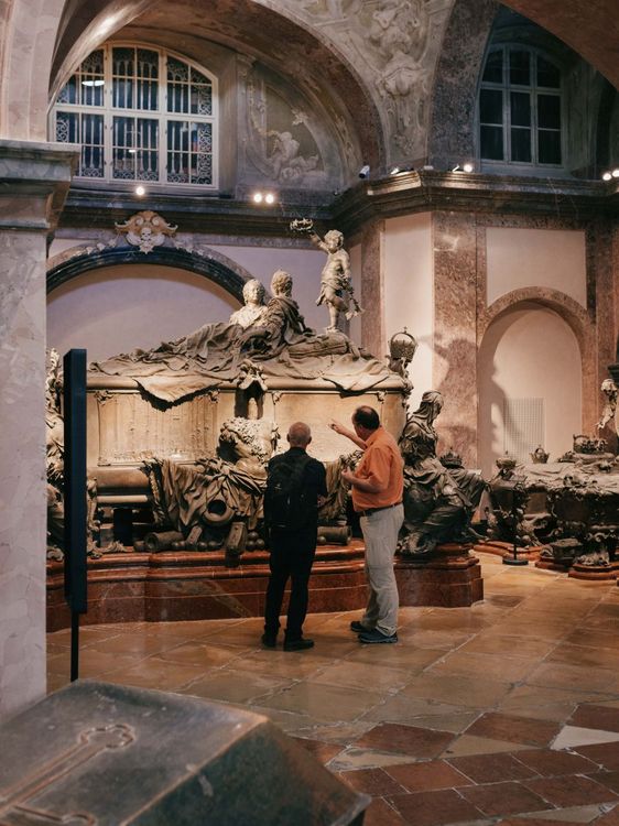 Kreuter zeigt einer anderen Person ein großes historisches Grabmal in einem Museumsraum. Die beiden sind von hinten zu sehen, wie sie das Grab betrachten.