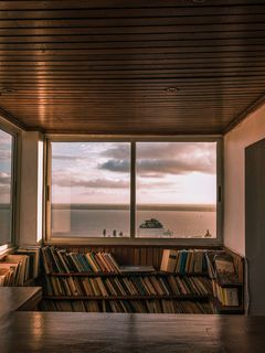Innenraum mit Holzdecke und Bücherregalen vor einem großen Fenster, das auf Meer, Himmel und ein Schiff blickt.