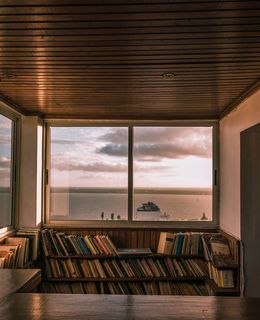 Innenraum mit Holzdecke und Bücherregalen vor einem großen Fenster, das auf Meer, Himmel und ein Schiff blickt.