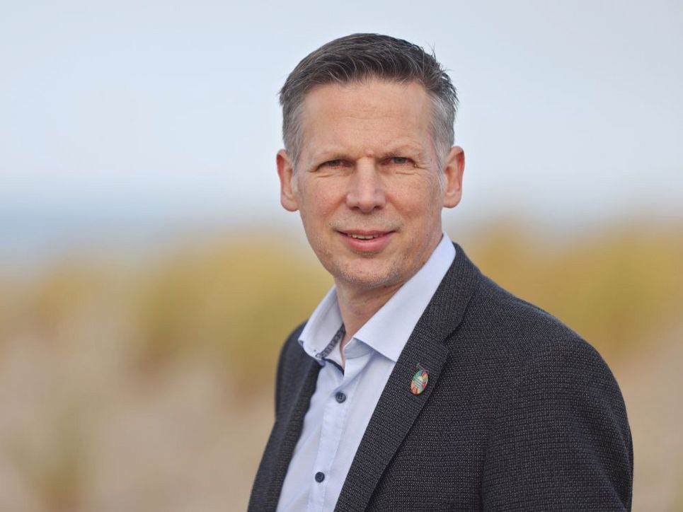Oliver Zielinski steht im Freien vor einer unscharfen Landschaft aus Dünen oder Grasflächen. Er trägt ein helles Hemd und ein dunkles Sakko mit einem kleinen bunten Anstecker und blickt leicht zur Seite in die Kamera.