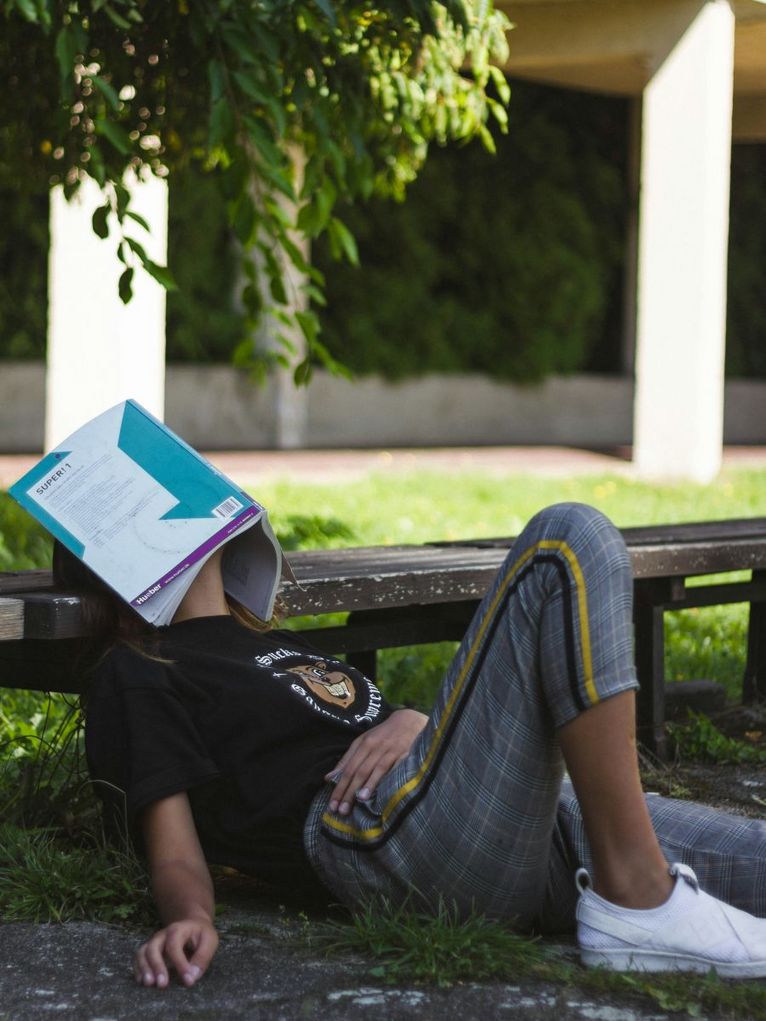 Eine Studentin sitzt mit einem aufgeklapptem Buch über dem Gesicht unter einem Baum.
