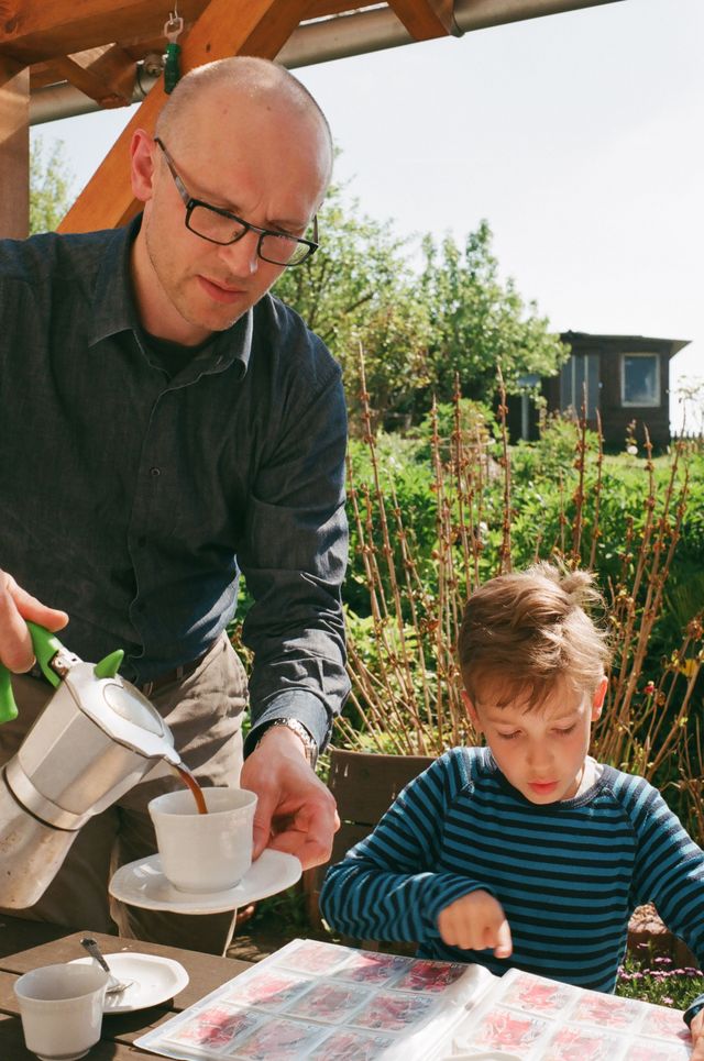 Der Vater gießt Kaffee ein, am Tisch auf der Veranda sitzt sein Sohn Kuba und schaut sich kleine Bildchen an.