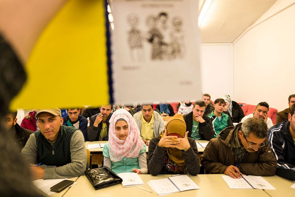 Viele Personen sitzen an Tischen, zum Teil Frauen, die Kopftuch tragen, manche schauen auf Aufgabenblätter.