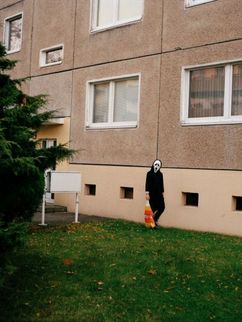 Eine Person mit Geistermaske und Norma-Tüte vor einem Plattenbau.