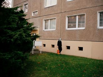 Eine Person mit Geistermaske und Norma-Tüte vor einem Plattenbau.