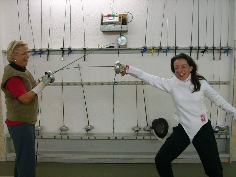 Annette Trabold in weißem Fecht-Anzug und in Fechtpose und eine weitere Frau mit Degen in der Hand. An der Wand hinter ihnen hängen viele Degen.