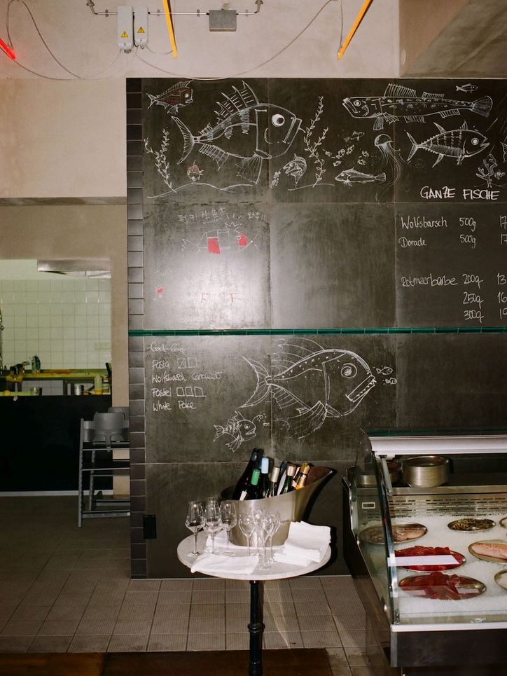 Tafel, die mit Fischen bemalt und mit Fischnamen beschriftet ist hinter einer Fischtheke und einem Tischchen auf dem eine eine Schale mit Weinflaschen und Weingläser stehen.