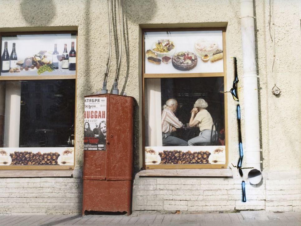 Durch das Fenster eines Osteuropäischen Lokals sieht man ein älteres Ehepaar an einem Tisch sitzen. Die Fenster des Lokals sind mit Fotos von Wein, Desserts und Kaffeebohnen dekoriert. Ein rostbrauner Stromkasten steht zwischen den Fenstern.