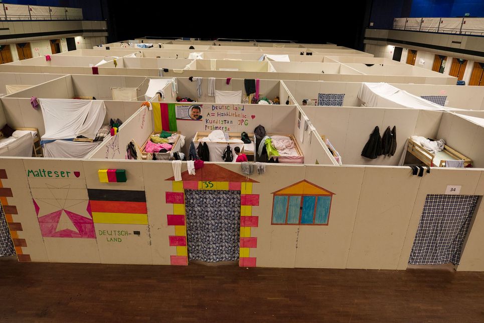 Blick in Räume einer Unterkunft für Geflüchtete, die durch Trennwände unterteilt und nach oben offen sind. An der Außenwand ist zu lesen: Malteser Deutschland. Daneben eine aufgemalte Tür.