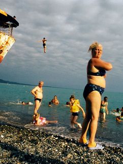Badende an einem steinigen Strand, ein altes Schiff wird als Sprungturm genutzt. Trotz bewölktem Himmel scheint die Sonne auf das türkisblaue Wasser.
