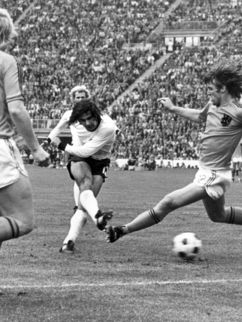 Schwarz-weiß Fotografie zeigt drei Fußballer beim Fußballspielen vor voll besetzten Rängen.