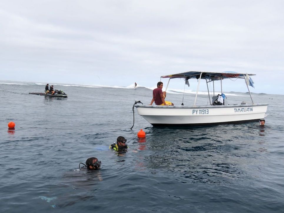 Zwei Personen mit Taucherbrillen im Wasser, ein Mann auf einem Boot, einer davor. Im Hintergrund Surfer und zwei Personen auf einem Motorsportboot.