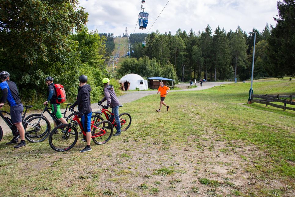 Hans G. Drexler läuft unter einer Seilbahn hindurch und wird von einer Gruppe E-Mountainbiker bestaunt.