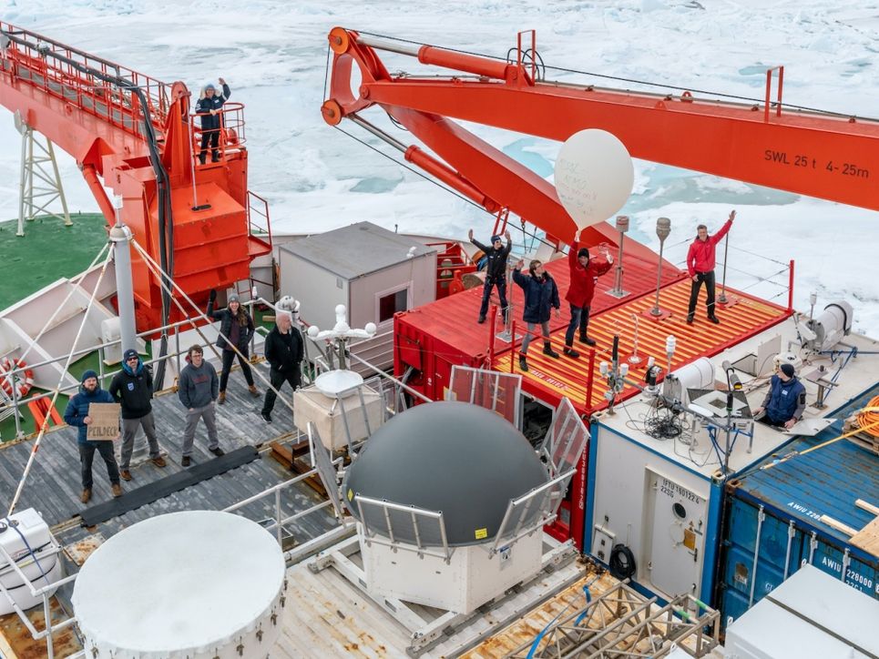 Deck des Schiffes mit Containern und orangenen Kranarmen, Personen stehen darauf, einige winken.