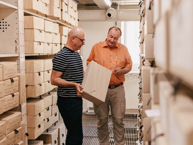 Kreuter und ein zweiter Mann stehen in einem Archivgang zwischen hohen Regalen mit beschrifteten Archivboxen. Einer hält eine Archivkassette in den Händen, während beide den Inhalt gemeinsam betrachten.