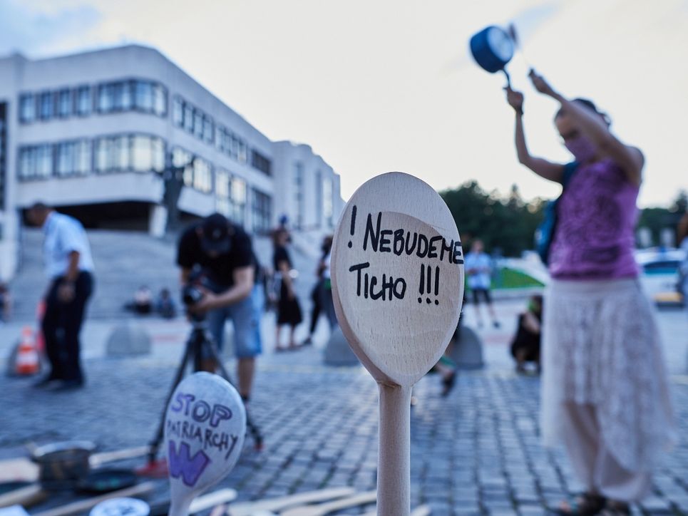 Eine Demonstrantin macht Lärm mit einem Topf und einem Kochlöffel. Auf dem Platz vor einem großen Gebäude sind weitere Personen, darunter ein Fotograf. Im Vordergrund ein Holzkochlöffel mit dem Motto »Nebudeme Ticho!« – »Wir werden nicht schweigen!«