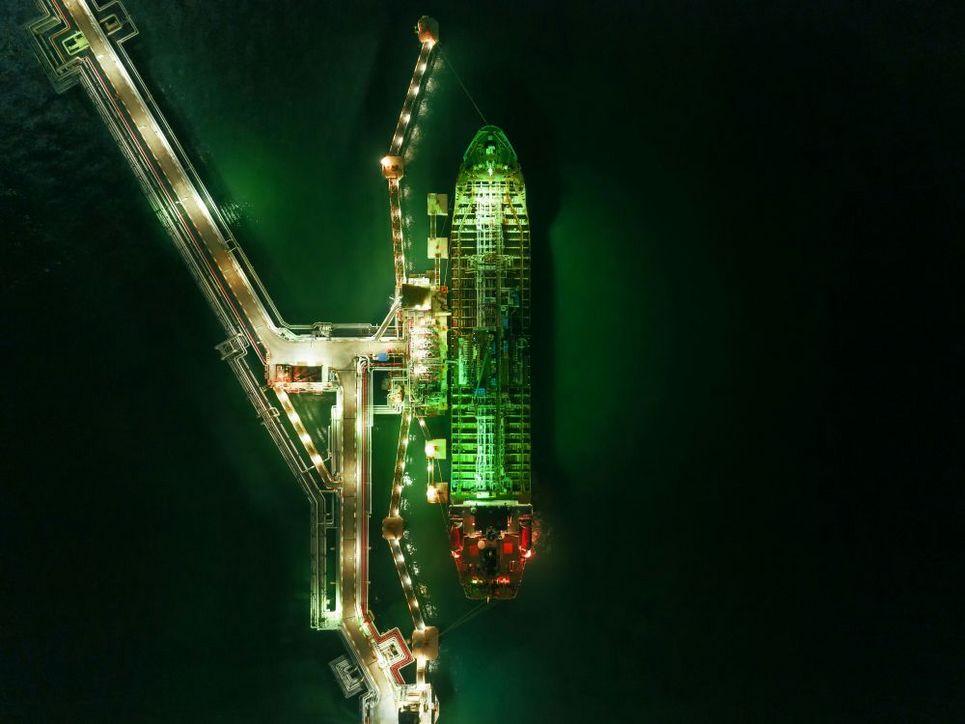 Eine nächtliche Luftaufnahme zeigt ein großes Schiff neben einem beleuchteten Pier. Das Schiff und die Hafenanlagen sind in grünes Licht getaucht und heben sich deutlich vom dunklen Wasser ab.
