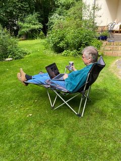 Ein Mann im Campingstuhl im Garten, mit Laptop, Getränk und Headset.