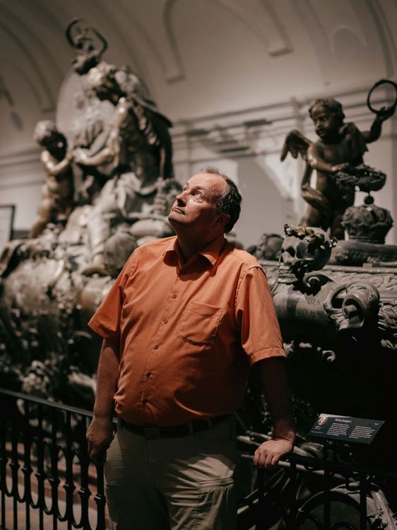 Kreuter in warmem Licht vor verschiedenen historischen Skulpturen. Kreuter trägt ein leuchtend orangenes Hemd mit kurzen Ärmeln. Und schaut zur Decke auf.