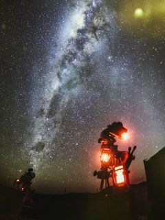 Im Hintergrund erstreckt sich die Milchstraße als helles Sternenband. Im unteren rechten Teil des Bildes ist die dunkle Silhouette einer technischen Konstruktion mit drei roten Lichtquellen zu sehen.