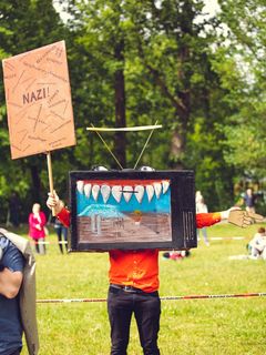 Person steht auf einer Wiese und hält ein Plakat mit der Aufschrift »Nazi!« und weiteren Aufschriften in die Höhe. Ihr Gesicht ist von einem weiteren Plakat verdeckt, auf dem unter anderem eine Reihe großer Zähne zu sehen ist, in der linken Hand hält sie eine Papierhand, die mit dem Zeigefinger von ihr aus gesehen nach links zeigt.