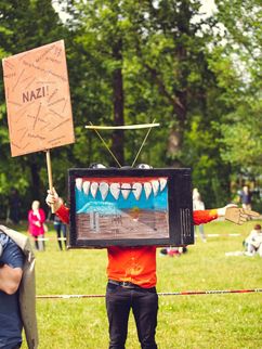 Person steht auf einer Wiese und hält ein Plakat mit der Aufschrift »Nazi!« und weiteren Aufschriften in die Höhe. Ihr Gesicht ist von einem weiteren Plakat verdeckt, auf dem unter anderem eine Reihe großer Zähne zu sehen ist, in der linken Hand hält sie eine Papierhand, die mit dem Zeigefinger von ihr aus gesehen nach links zeigt.