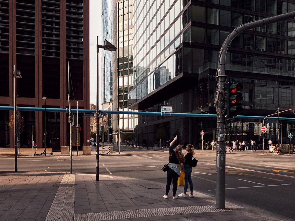 Zwei junge Frauen an einer Kreuzung, eine zeigt nach oben zu einem Hochhaus im Frankfurter Bankenviertel.