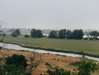 Weite Flusslandschaft an einem verregneten Tag, im Hintergrund Bäume.
