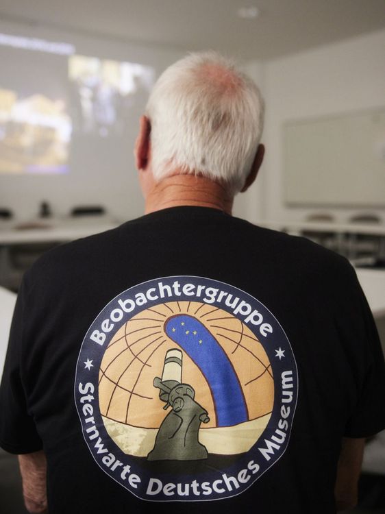 Rückansicht einer Person in einem Raum, die ein schwarzes T-Shirt mit einem großen Emblem und der Aufschrift Beobachtergruppe Deutsches Museum trägt.