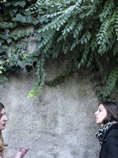 Zwei junge Frauen im Gespräch. Sie stehen vor einer grün bewachsenen Mauer.