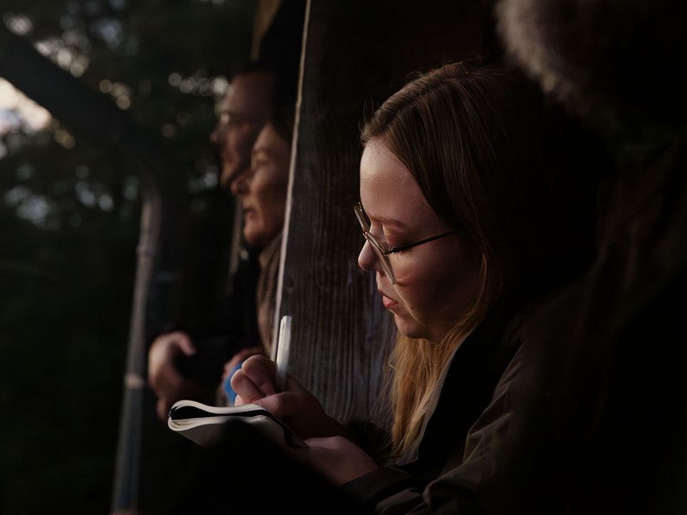 Blonde Frau mit Brille im Vordergrund schreibt in ein Notizbuch. Im Hintergrund schauen ein Mann und eine Frau in die Ferne.