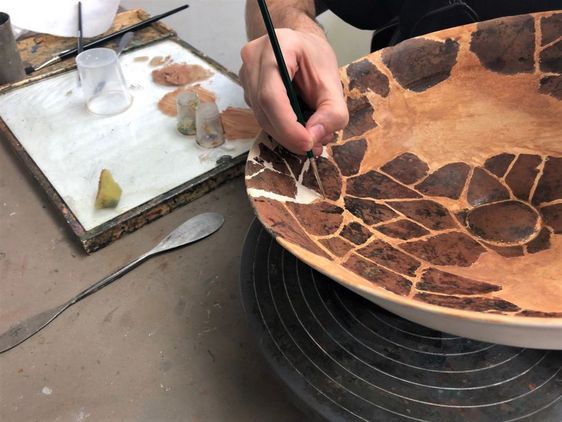 Eine getöpferte Schale mit mosaikartigem Muster wird innen mit einem feinen Pinsel bemalt.