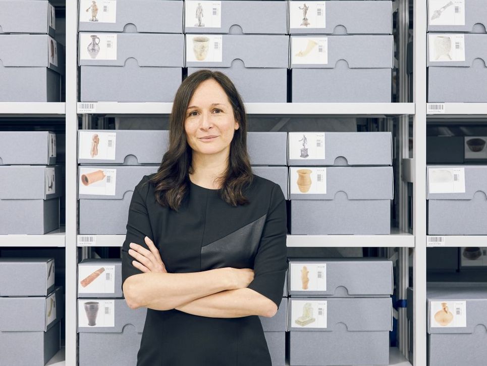 Alexandra W. Busch vor einem Regal mit grauen Kisten, auf denen Fotos von Archäologischen Objekten kleben.