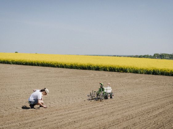 Felix Erben in der Hocke auf dem Acker hinter dem Roboter. Im angrenzenden Feld blüht der Raps.