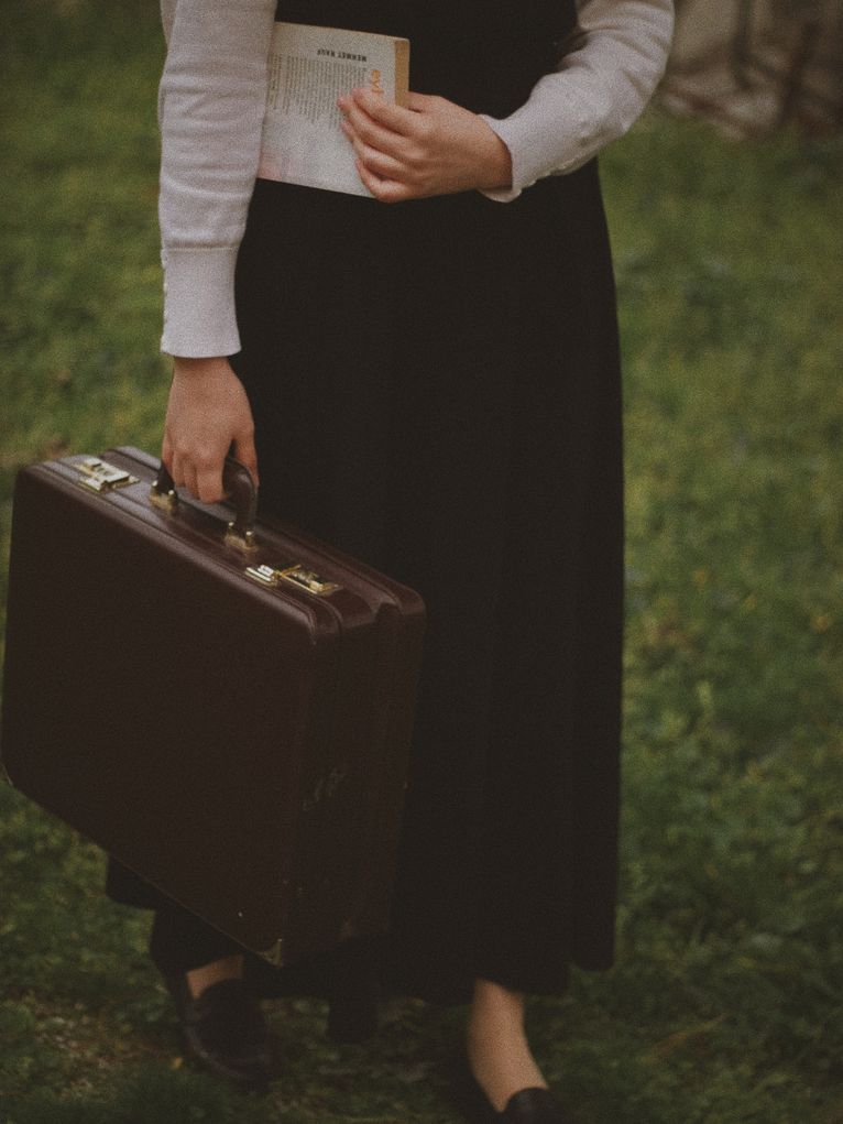 Eine Person, die einen Rock trägt und einen Koffer in der Hand hält und ein Buch unter den Arm geklemmt hat. Es sind nur von den Füßen bis zur Hälfte des Oberkörpers zu sehen.