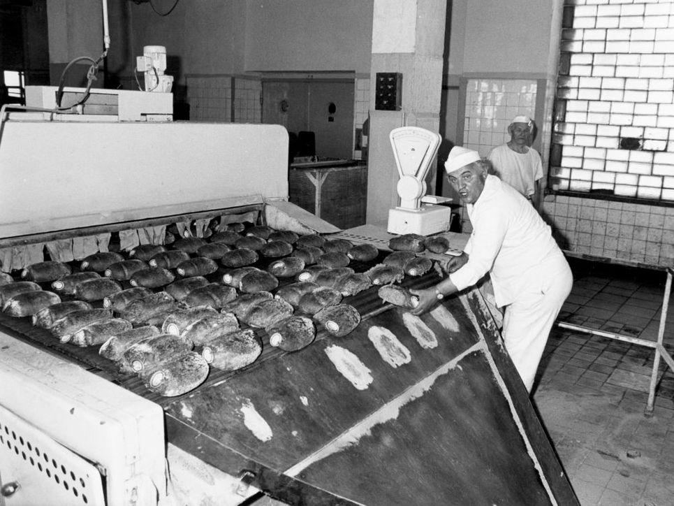 In einer Bäckerei arbeitet ein Mann an einer Maschine, auf der zahlreiche Brote liegen. Ein weiterer Mitarbeiter steht im Hintergrund, während die Schwarzweißaufnahme den Produktionsprozess in der Backstube zeigt.